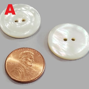 May include: Two white, round buttons with two holes each, next to a copper penny. The buttons have a pearlescent sheen and a slightly raised edge. The penny is dated 2017.