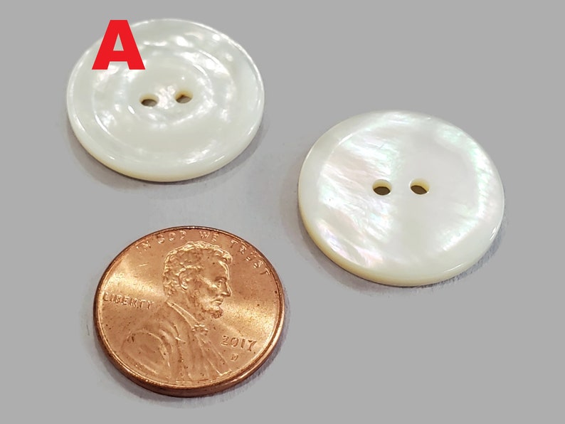May include: Two white, round buttons with two holes each, next to a copper penny. The buttons have a pearlescent sheen and a slightly raised edge. The penny is dated 2017.