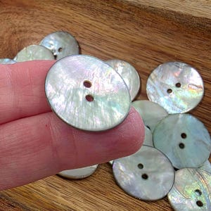 May include: A collection of iridescent, oval-shaped buttons with two holes each. The buttons have a pearlescent sheen and are made of a light-colored material. The buttons are scattered on a wooden surface.