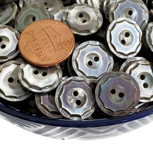 May include: A bowl filled with a large quantity of iridescent, mother-of-pearl buttons. The buttons are two-hole, round, and have a scalloped edge. A US penny is included for scale.