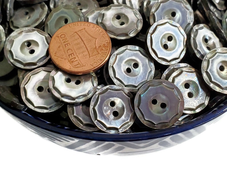 May include: A bowl filled with a large quantity of iridescent, mother-of-pearl buttons. The buttons are two-hole, round, and have a scalloped edge. A US penny is included for scale.