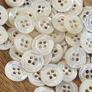 Può includere: Una collezione di bottoni rotondi color bianco sporco con una finitura perlata. Ogni bottone presenta un centro incassato con quattro fori e un bordo ottagonale rialzato. I bottoni sono disposti in un mucchio denso e sovrapposto, mostrando le loro superfici lisce e riflettenti e un design coerente in tutto il gruppo.