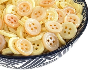 May include: A close-up shot of a bowl filled with a variety of round, cream-colored buttons. Some buttons have a peach-colored center. The buttons have four holes for sewing. The bowl has a blue and white patterned rim.