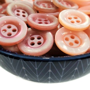 Op de afbeelding: Een kom gevuld met roze en witte iriserende knopen. De knopen hebben vier gaten en zijn in een willekeurig patroon gerangschikt.