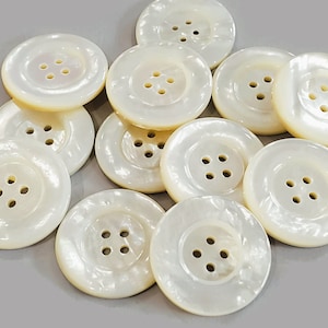 Peut inclure: Un ensemble de 12 boutons en nacre blanche, chacun avec quatre trous. Les boutons sont ronds et ont un bord légèrement surélevé.
