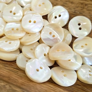 May include: A collection of iridescent, off-white buttons with two holes each. The buttons vary in shape, including round, teardrop, and irregular forms. The buttons have a pearlescent sheen, reflecting light. The buttons are scattered on a wooden surface.