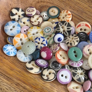 May include: A collection of assorted vintage buttons in various colors and designs. The buttons are made of different materials and feature a range of patterns, including stripes, dots, and floral motifs. The buttons are in a wooden bowl.