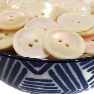 May include: A bowl filled with white iridescent buttons. The buttons have two holes and are arranged in a random pattern. The bowl is blue and white with a geometric design.