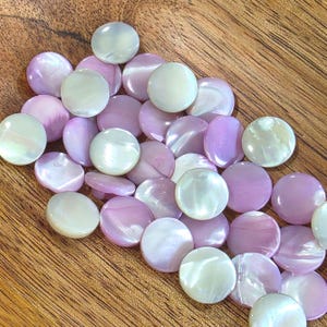 May include: A collection of small, round, flat buttons featuring a smooth, iridescent finish. The buttons alternate between a soft, pale lavender purple and a creamy, pearlescent white, each displaying a natural, glossy sheen across the surface.