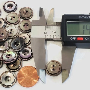 May include: A pile of 18 iridescent mother of pearl buttons with a scalloped edge and two holes. The buttons are 1.437 inches in diameter. A penny is included for scale.