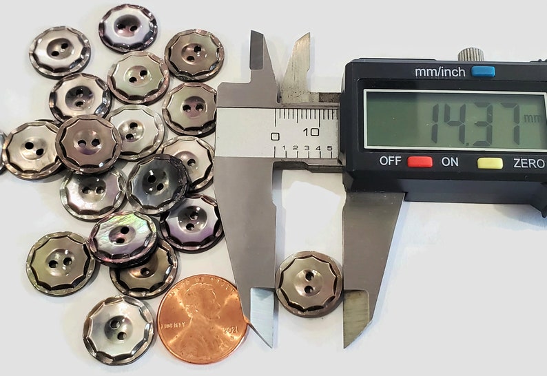 May include: A pile of 18 iridescent mother of pearl buttons with a scalloped edge and two holes. The buttons are 1.437 inches in diameter. A penny is included for scale.