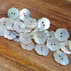 May include: A collection of iridescent, flower-shaped buttons with two holes each. The buttons are white with a pearlescent sheen, scattered on a wooden surface. The buttons have a scalloped edge, and the wood grain is visible.