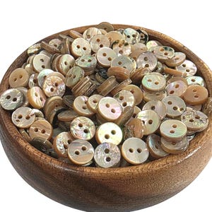 May include: A large collection of round, two-hole buttons made from iridescent mother-of-pearl shell, featuring natural variations in tan, brown, and cream tones, all contained within a smooth, circular wooden bowl with a visible grain pattern.