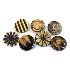 Puede incluir: Una colección de ocho botones vintage con varios diseños. Los botones presentan una gama de colores, incluyendo negro, crema y marrón. Algunos botones tienen patrones de rayas, florales o geométricos. Los botones son redondos y parecen ser de plástico.