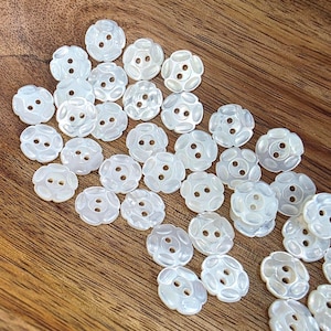 May include: A collection of white, flower-shaped buttons with two holes each. The buttons have a pearlescent sheen and are scattered on a wooden surface. These decorative buttons are ideal for sewing and craft projects.