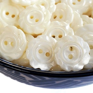 Puede incluir: Primer plano de botones blancos en forma de flor, llenando un cuenco azul y blanco. Cada botón tiene dos agujeros y un borde festoneado. El conjunto sugiere un tema vintage o artesanal, ideal para costura.