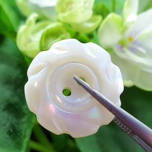 以下が含まれることがあります： 波打つ縁取りと2つの穴のある、白く虹色に輝く真珠貝のボタン。ピンセットで挟まれている。