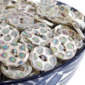 Puede incluir: Un bol lleno de botones blancos iridiscentes con un patrón de lunares negros y blancos. Los botones tienen un brillo iridiscente arcoíris.