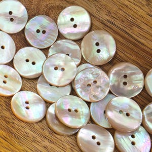 May include: A collection of iridescent, round buttons with two or four holes. The buttons have a pearlescent sheen with hints of pink, green, and yellow. The buttons are scattered on a wooden surface.