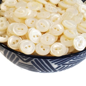 Può includere: Primo piano di una ciotola piena di numerosi bottoni bianco-avorio perlati. I bottoni sono rotondi con due fori e un bordo decorativo a forma di corda. La ciotola ha un motivo geometrico blu e bianco.