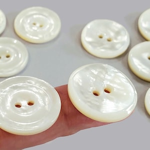 May include: A collection of round, off-white buttons with two holes each. The buttons have a pearlescent sheen and a slightly domed shape. They are arranged on a light gray surface, suggesting a craft or sewing project.