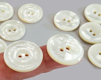 May include: A collection of round, off-white buttons with two holes each. The buttons have a pearlescent sheen and a slightly domed shape. They are arranged on a light gray surface, suggesting a craft or sewing project.
