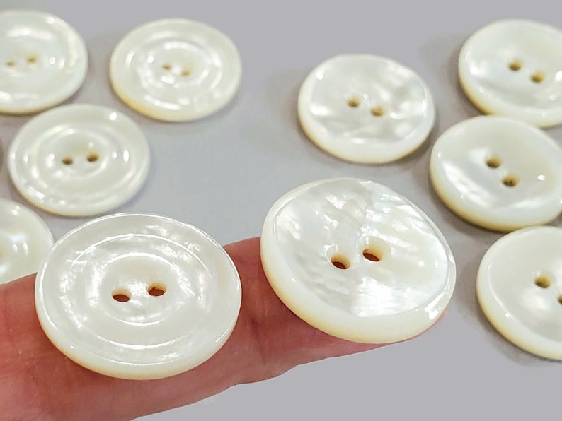 May include: A collection of round, off-white buttons with two holes each. The buttons have a pearlescent sheen and a slightly domed shape. They are arranged on a light gray surface, suggesting a craft or sewing project.