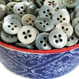 Op de afbeelding: Een kom gevuld met een grote hoeveelheid grijze en witte parelmoerknopen. De knopen hebben vier gaten en bevinden zich in een kom met een blauw-wit patroon en een rode rand.