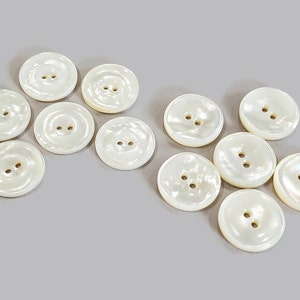 May include: A collection of round, off-white buttons with two holes each. The buttons have a pearlescent sheen and are scattered on a light gray surface. These buttons are suitable for sewing and craft projects.