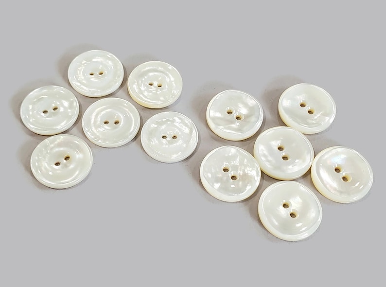 May include: A collection of round, off-white buttons with two holes each. The buttons have a pearlescent sheen and are scattered on a light gray surface. These buttons are suitable for sewing and craft projects.