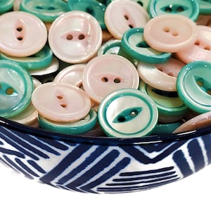 Op de afbeelding: Een kom gevuld met verschillende twee-gaats knopen in roze en turquoise tinten. De knopen zijn gemaakt van parelmoer en hebben een gladde, glanzende afwerking.