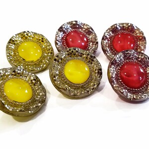 May include: A collection of six decorative buttons. The buttons have a textured, silver-colored rim and a central, rounded gem. Three buttons have yellow gems, and three have red gems. The buttons are on a white background.