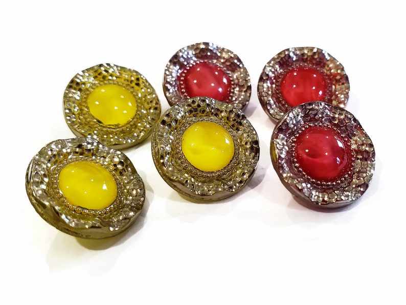 May include: A collection of six decorative buttons. The buttons have a textured, silver-colored rim and a central, rounded gem. Three buttons have yellow gems, and three have red gems. The buttons are on a white background.
