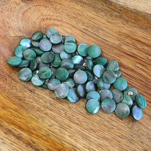 May include: A collection of numerous round, flat buttons featuring a smooth, iridescent surface with swirling shades of deep forest green, teal, and muted gray. Each button has a polished finish that reflects light, creating a varied color depth.