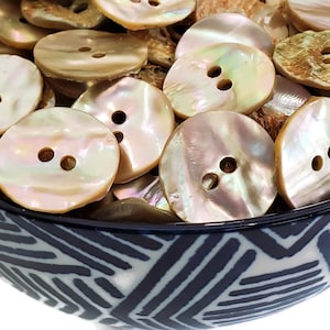 Puede incluir: Colección de botones redondos iridiscentes con dos agujeros, en tonos crema, beige y dorado. Los botones están en un cuenco decorativo con un patrón geométrico azul y blanco.