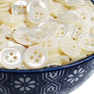 May include: A bowl filled with white mother of pearl buttons. The buttons have four holes and a smooth, shiny finish.
