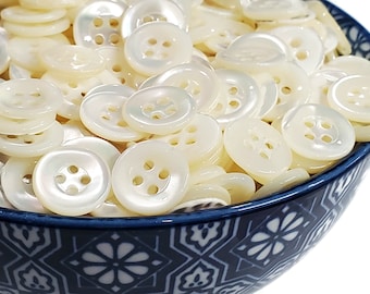 May include: A bowl filled with white mother of pearl buttons. The buttons have four holes and a smooth, shiny finish.