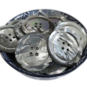 May include: A collection of large, iridescent gray buttons with four holes each, displayed in a blue and white patterned bowl. The buttons have a pearlescent sheen, reflecting light. Ideal for sewing and craft projects.