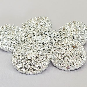May include: A silver-toned, flower-shaped embellishment covered in small, sparkling rhinestones. The five-petal design is set against a light gray background, creating a dazzling effect. This decorative piece could be used for crafts or fashion.