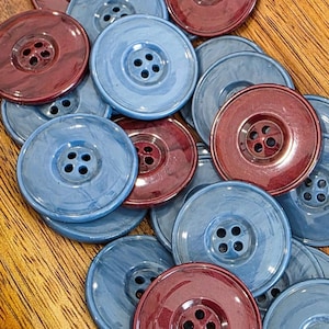 May include: A collection of large, round buttons in shades of blue and burgundy. Each button has four holes for sewing and a glossy finish. The buttons are scattered on a wooden surface, creating a textured and colorful display.