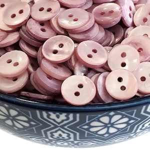 May include: A bowl filled with pink iridescent shell buttons. The buttons have two holes and are in a variety of sizes.
