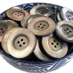 May include: A collection of iridescent, pearlescent buttons with four holes, in a variety of sizes, are piled in a blue and white patterned bowl. The buttons have a subtle, reflective sheen.