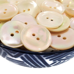 May include: A bowl filled with a variety of white pearl buttons with two holes each. The buttons are smooth and have a slight iridescent sheen.