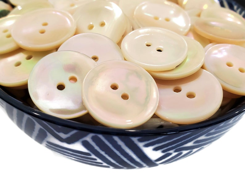 May include: A bowl filled with a variety of white pearl buttons with two holes each. The buttons are smooth and have a slight iridescent sheen.