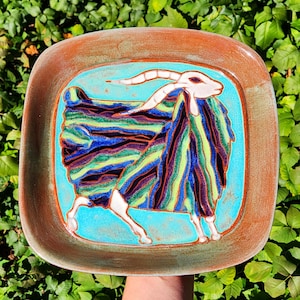 May include: A square ceramic plate with rounded corners featuring a hand-painted goat illustration. The goat has a white head and legs against a bright turquoise background, with its body filled with vertical stripes of blue, green, and purple.