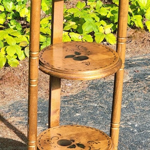 Puede incluir: Un estante para plantas de madera de tres niveles con acabado marrón claro. Cada estante circular presenta un diseño floral oscuro. El soporte tiene una parte superior e inferior decorativas y mide aproximadamente 102 cm de alto.