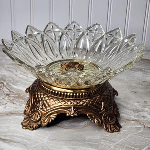 May include: A clear glass bowl with a flower petal design sits atop a decorative bronze-colored metal base. The base has ornate detailing and a round, textured center. The bowl is likely for decorative purposes.