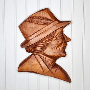 May include: A carved wooden wall hanging depicting a profile of a person wearing a hat with a feather. The wood has a warm brown tone, and the carving details the person's features and clothing. The background is a white vertical-slat wall.