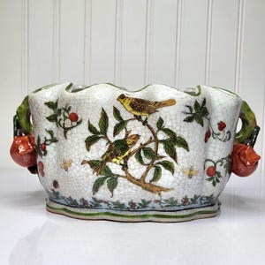 Peut inclure: Un cache-pot en céramique blanche avec un bord festonné et une finition craquelée. L'extérieur présente un motif peint à la main de deux oiseaux sur des branches, entourés de feuilles vertes, de baies rouges et de petites abeilles. Deux anses en forme de fruit rouge.