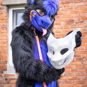 Könnte beinhalten: Ein Ganzkörper-Fursuit aus schwarzem und violettem Kunstfell mit orangefarbenen Akzenten und einer rosa Zunge, der eine schlichte, unbemalte, tierförmige Maske hält. Der Anzug hat eine violette Schnauze, orange Ohrspitzen und einen orangefarbenen Riemen.